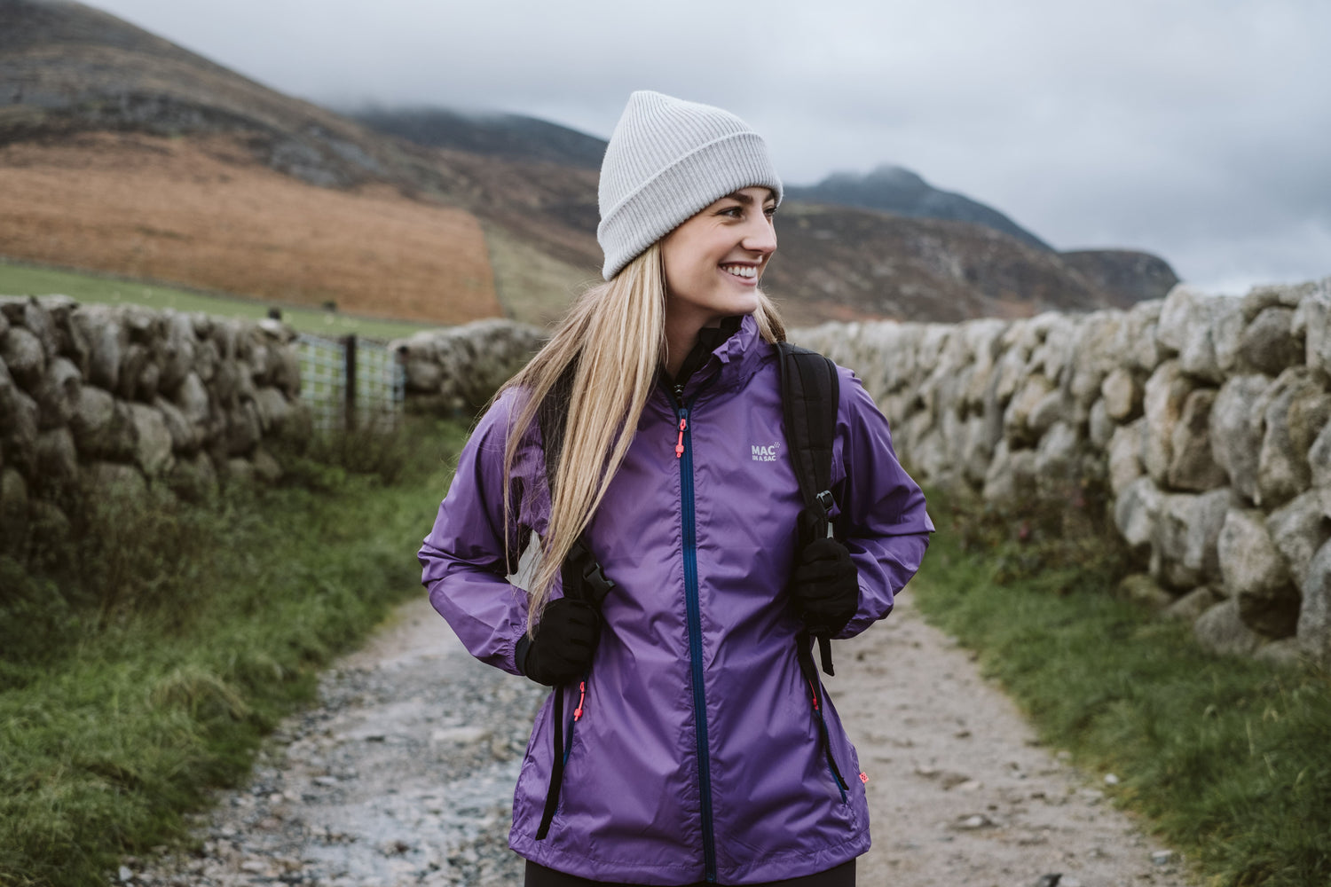 Woman in a purple jacket and light grey beanie standing on a path with stone walls and mountains in the background.