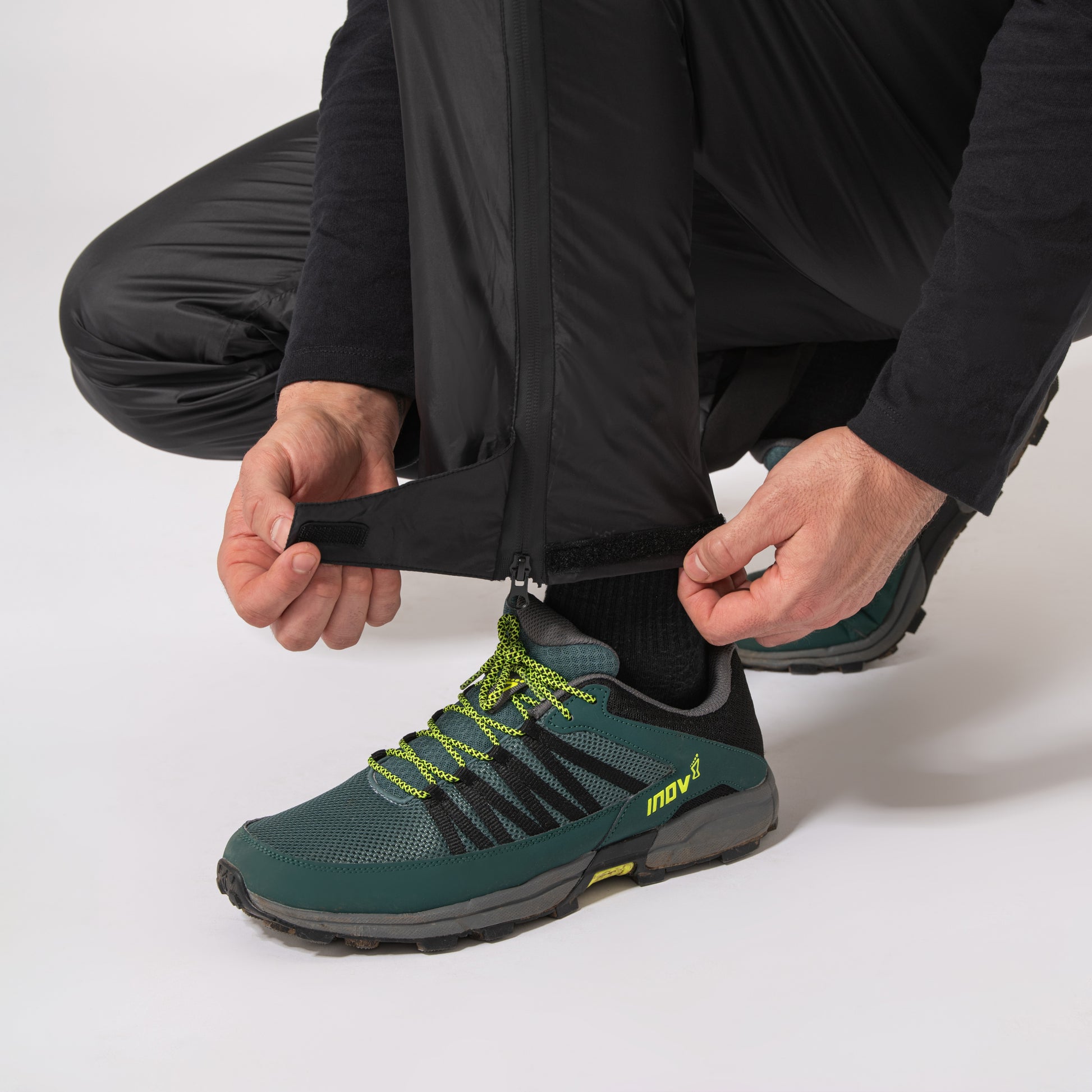 black waterproof overtrousers displaying the adjustable velcro straps while kneeling down with a white background